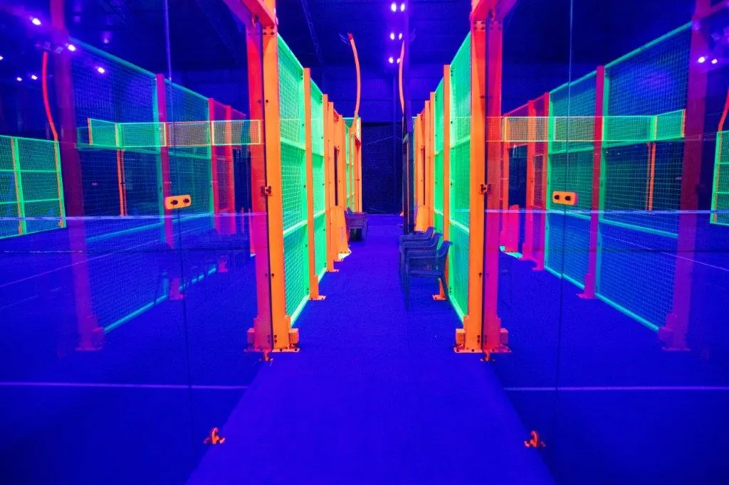 Walkway between two glow padel courts with neon green and orange fences under blue UV lighting
