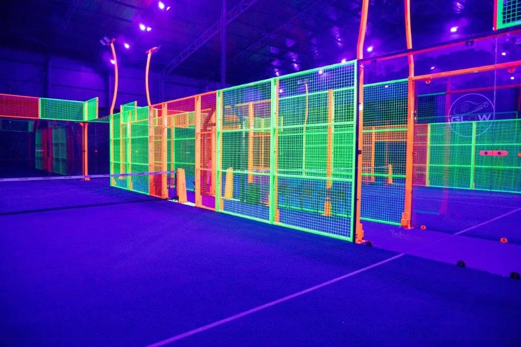 Glow padel court seen from the baseline, with the net, neon fence panels and UV lighting in an indoor venue