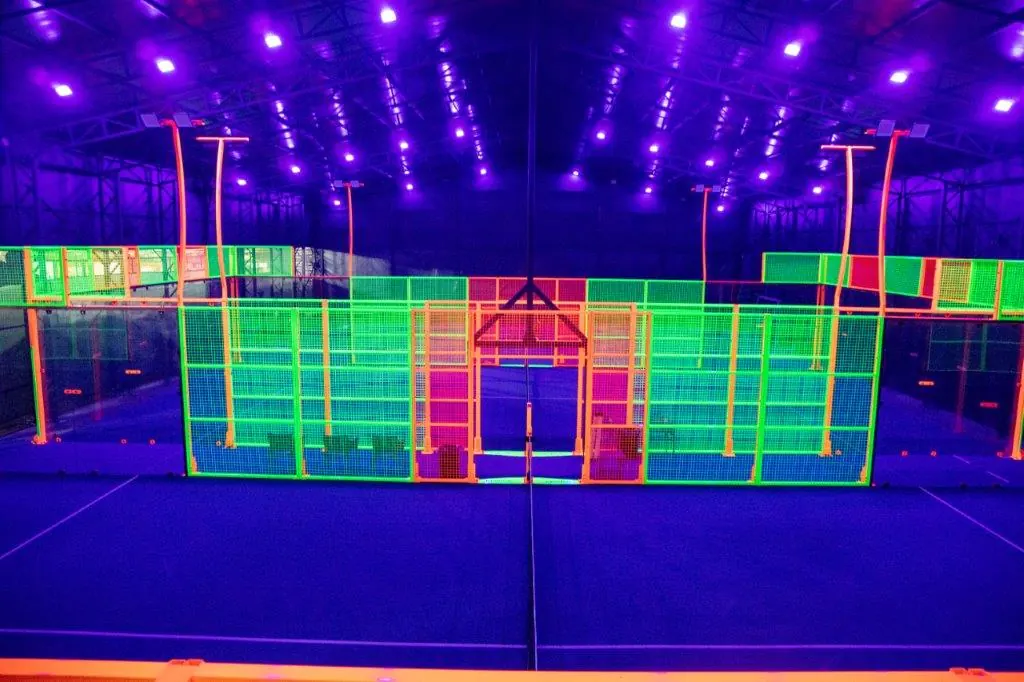 Indoor glow padel court lit by UV lights, with neon green and orange fences glowing against a deep blue floor